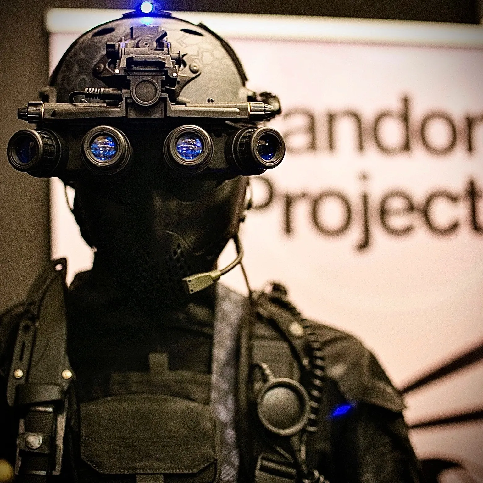 A figure in night vision goggles in front of a lit backdrop at an event